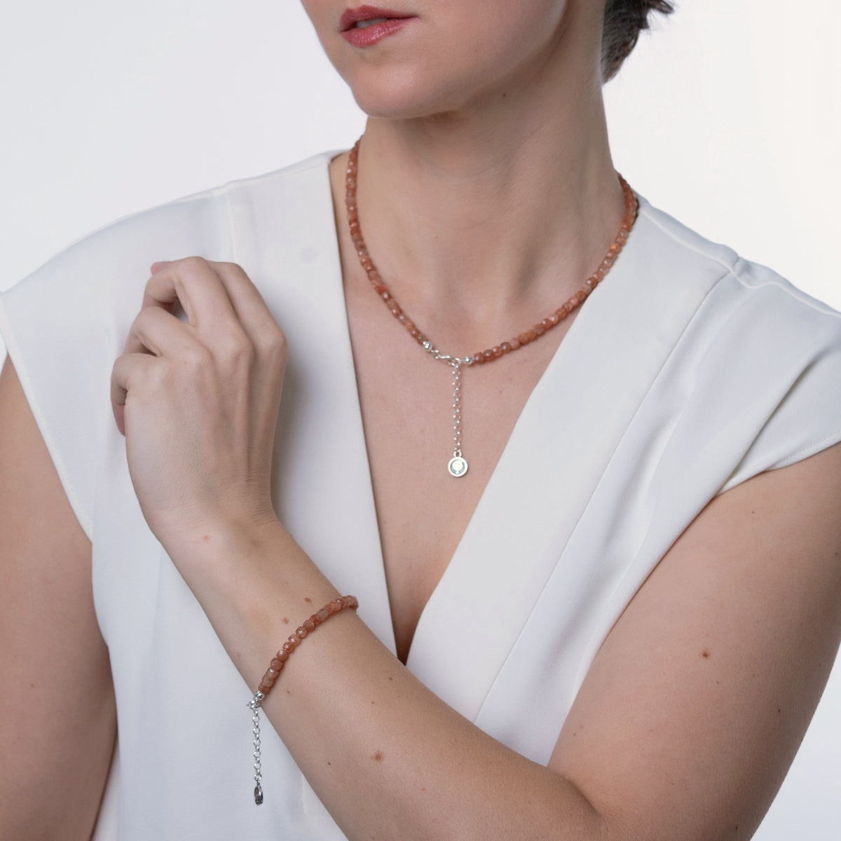 A woman wearing a beaded necklace and matching bracelet in a warm terracotta color with silver-toned chain details and a round disc charm, styled with a white sleeveless top against a light gray background.