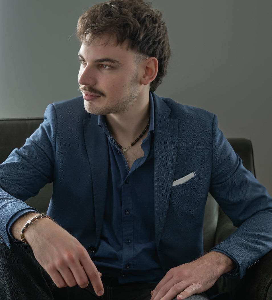 Man in a blue suit with amber necklace and bracelet is sitting on a couch in a neutral setting