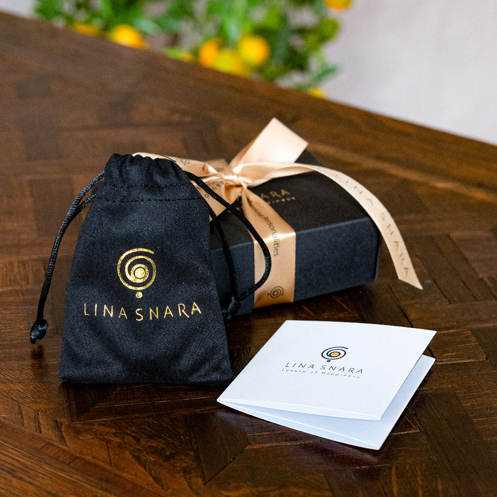 Lina Snara jewelry packaging with black drawstring bag, box, and card on a wooden surface.